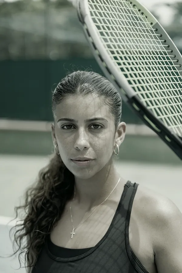 A coach with her dark hair tied back looks into the camera, a racket over her shoulder.
