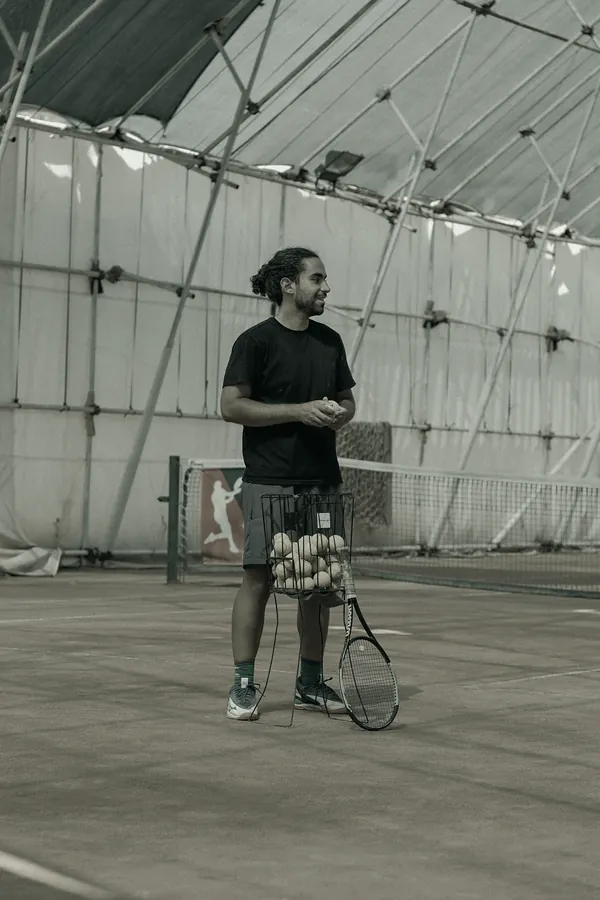 A coach in a black T-shirt stands on a clay court beside a basket of balls under a white tent.