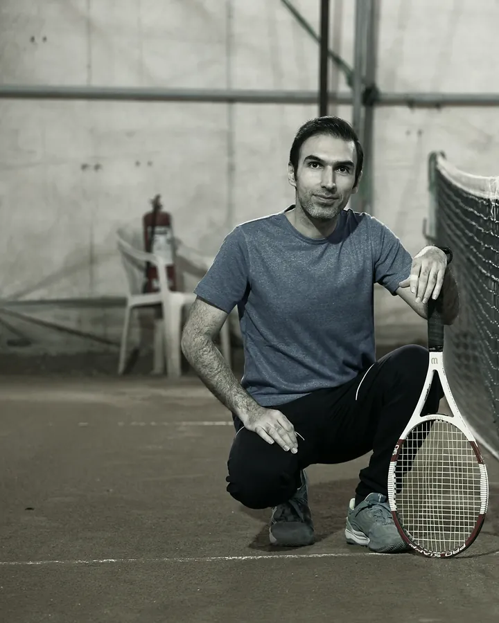 A smiling coach crouches by the net on a clay court, holding his racket.