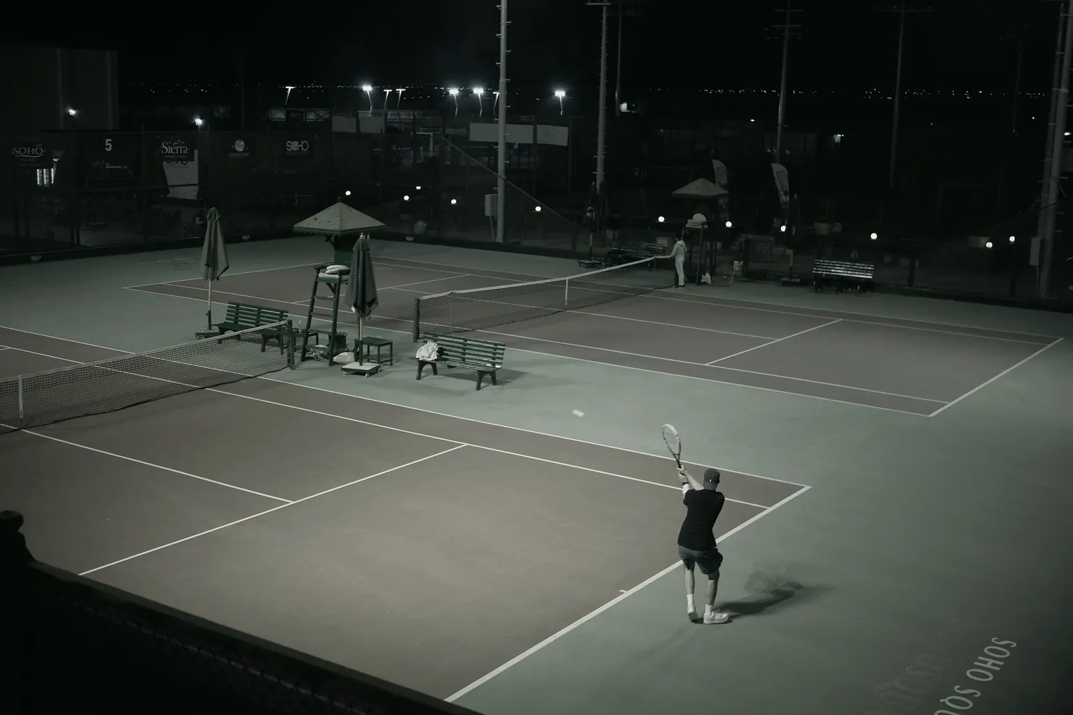 Floodlit courts at night, a clay court and a hard court side by side, one player serving.