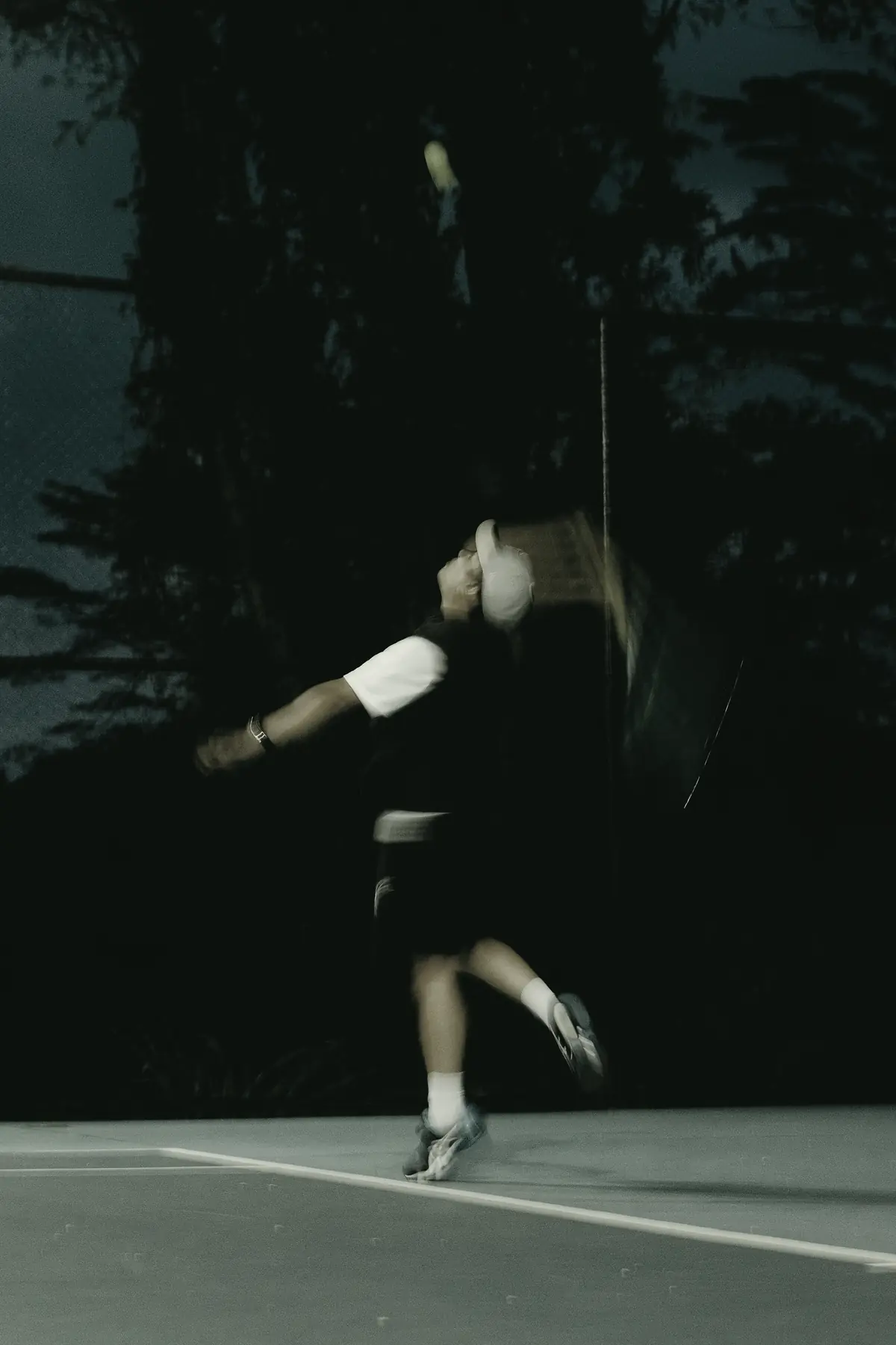 At dusk a player in a white cap swings through a serve, the racket a blur against dark trees.