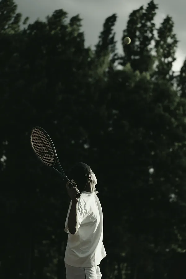 A player in white looks up at the ball before a serve, tall dark trees behind him.