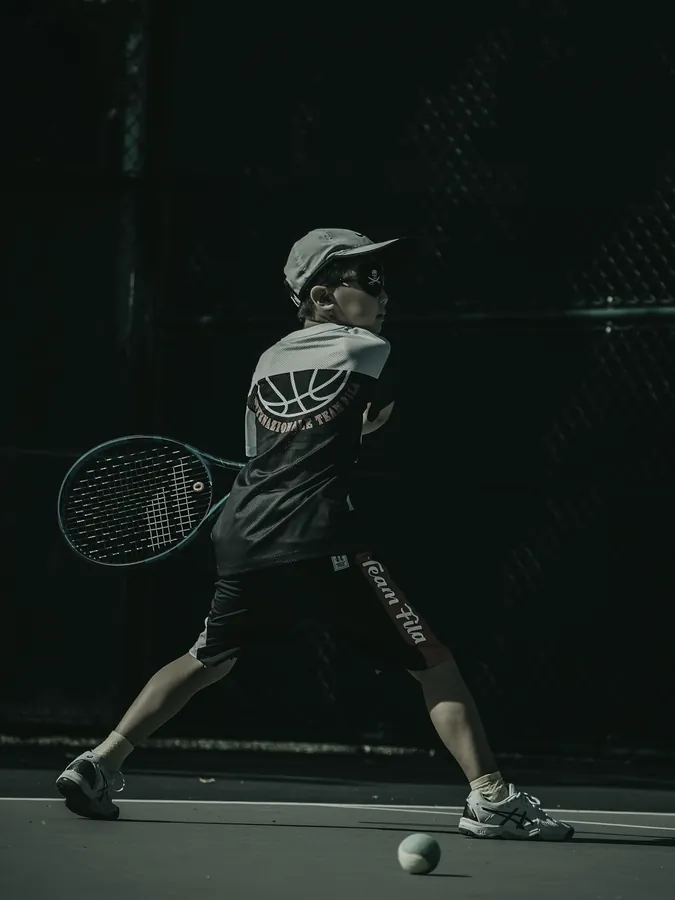 A boy in a cap and sunglasses swings a forehand in front of a dark green fence.