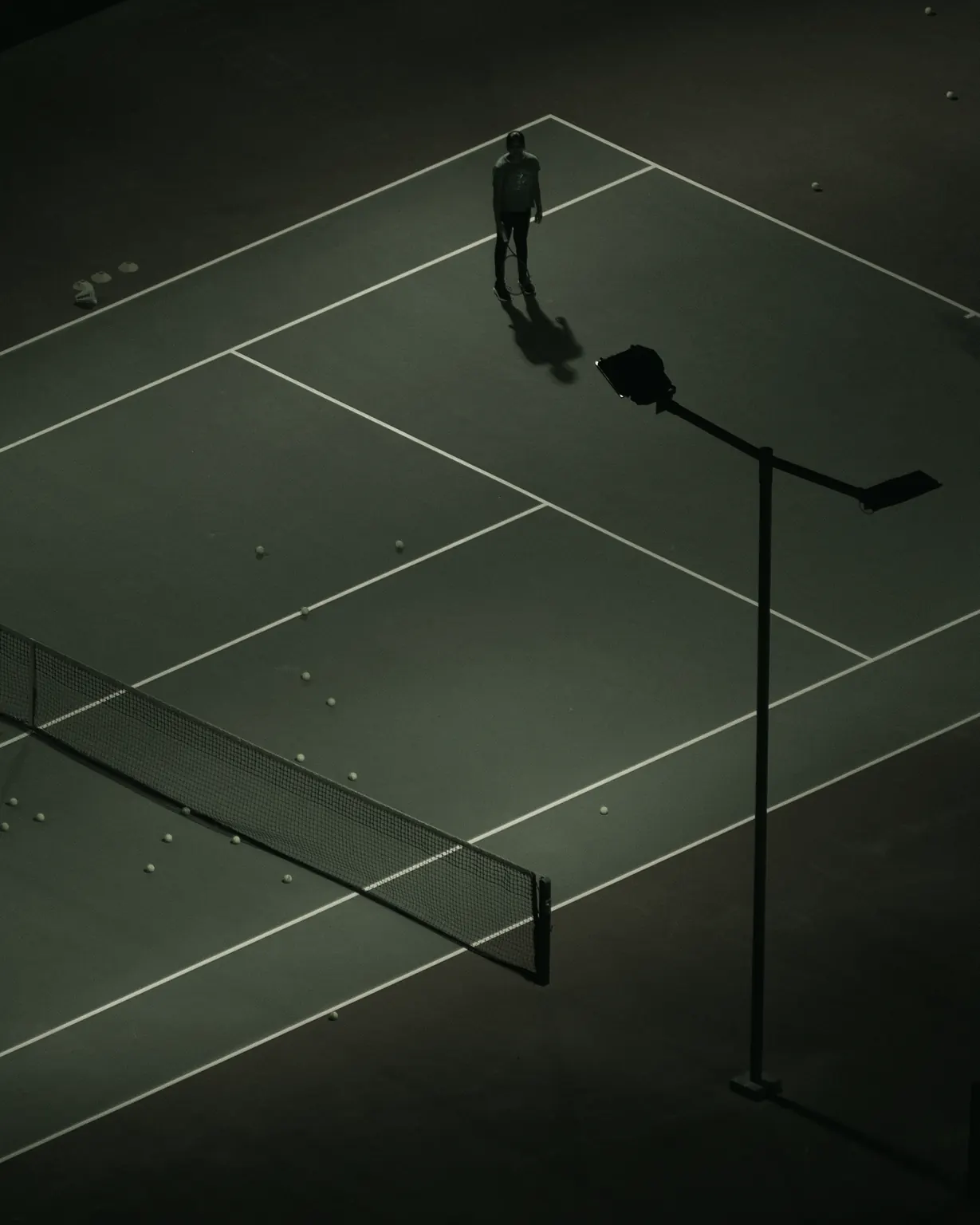 Seen from above, a coach stands on a dark green court scattered with balls, a floodlight beside the net.